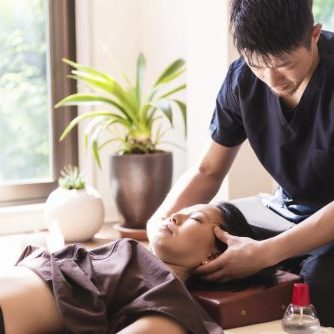 Young woman, Japanese ethnicity, at the acupuncture treatment. Young doctor acupuncturist massaging her face before starting the treatment. She is lying,  at bright window.